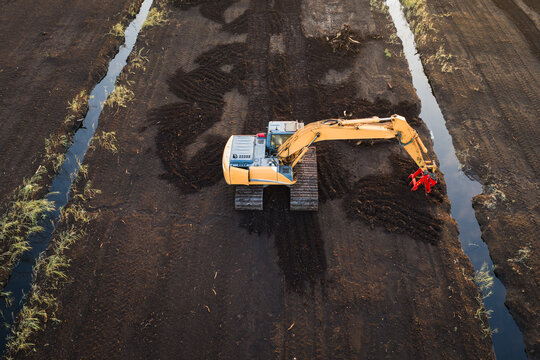 An aerial view from a drone shows a yellow tracked excavator working on a dark peat bog, digging drainage channels with a red attachment.