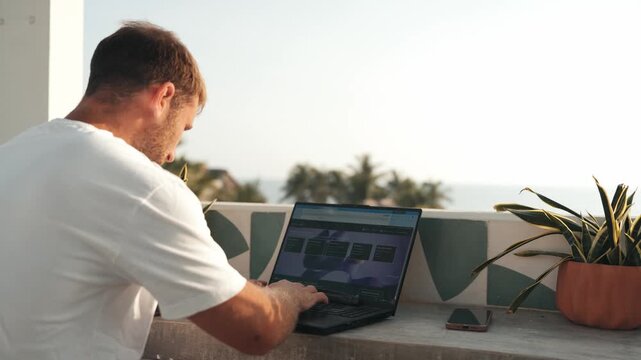 Young man typing on a laptop computer while enjoying a beautiful ocean view from a sunny balcony