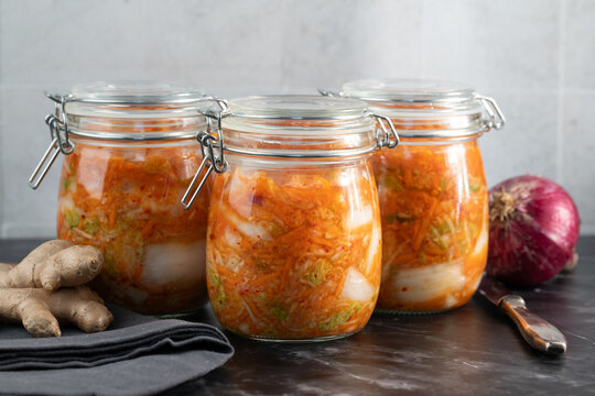 Freshly prepared kimchi is being poured into jars in the kitchen. In the background are an onion and ginger.