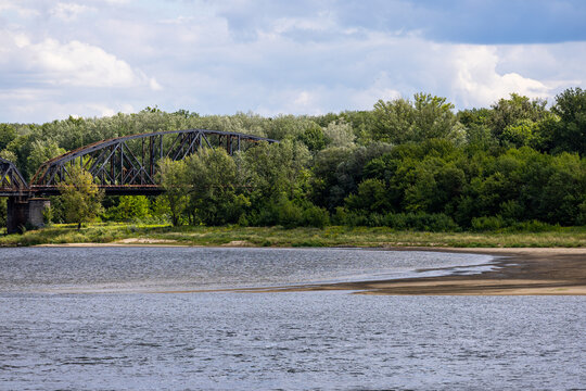 Steel truss bridge stands behind a calm river with sandbars near the shore. Dense trees and vegetation line the riverbank. Simple natural setting supports themes of transport.