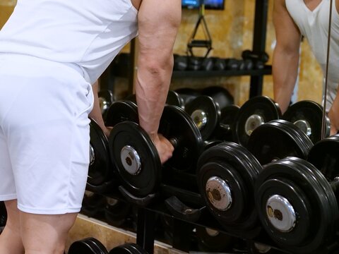 Close-up of muscular man grabbing heavy black dumbbell from weight rack in gym mirror reflection.