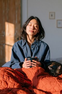 Woman enjoying morning coffee in bed with eyes closed wearing blue shirt and orange quilt for cozy bedroom background and calm lifestyle imagery