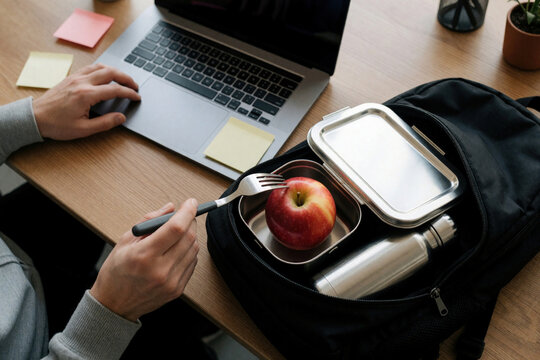 Person works on laptop at desk. Nearby, an open backpack contains lunchbox, apple, and water bottle. Healthy eating, remote work and study setup shown.