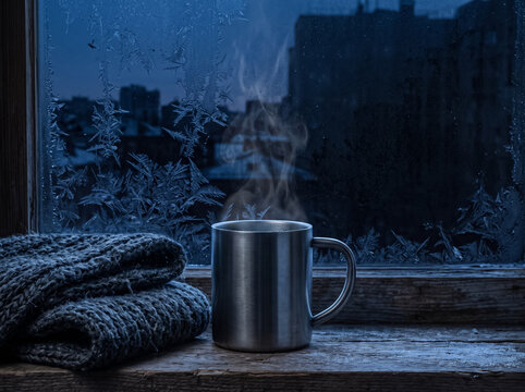 a metal mug with hot steam standing on a wooden windowsill next to a folded blanket against a frozen window background