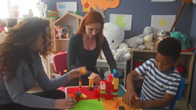 Women and boy building colorful interlocking toy blocks with hands at a playroom table inside a building; focused learning.