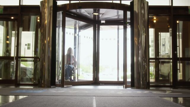 Young adult Caucasian woman with disability in wheelchair entering modern office building through revolving door. Footage is ideal for corporate diversity and accessibility campaigns