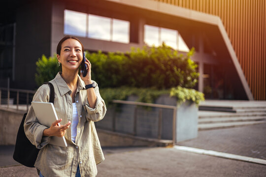 Smiling Young Student Woman Talking on Phone While Carrying Laptop Outside Modern Campus Building