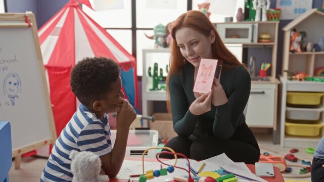 Woman teacher shows and points to a flashcard while kneeling with child in a colorful preschool classroom; early literacy curiosity.