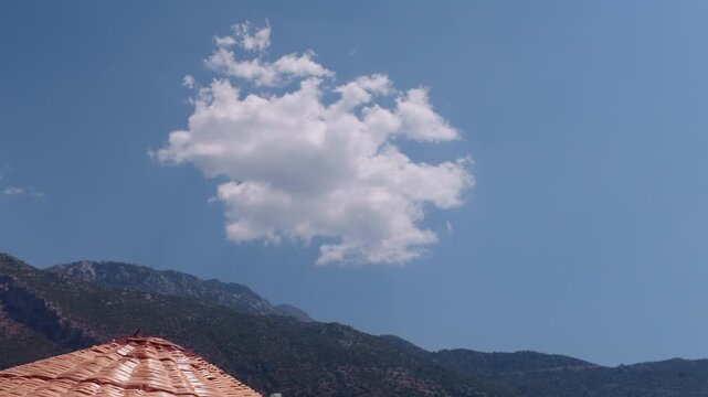 View from under a straw beach umbrella at a fluffy white cloud over Babadağ mountain, with a red paraglider later flying into the clear blue sky, Oludeniz, Turkey.