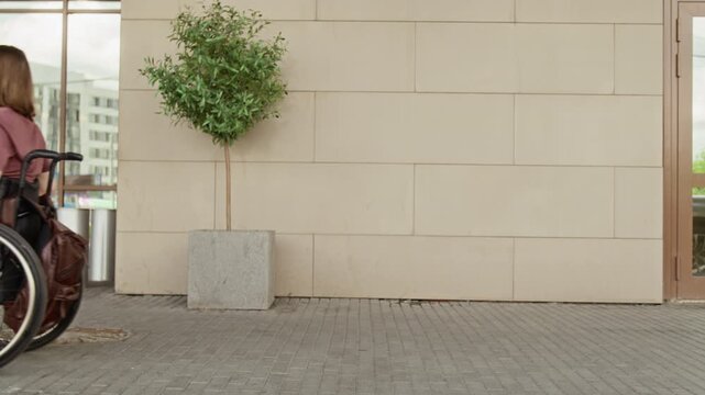 Young adult Caucasian woman with disability riding wheelchair along modern building wall, turning around. Footage is ideal for social diversity and inclusion campaigns