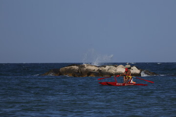 Lifeguard rowing a traditional red rescue boat past a breakwater on the Adriatic coast © Ionel