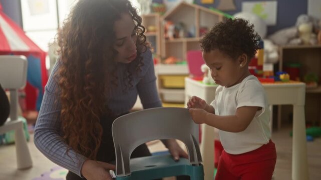 Woman bends to help a toddler grip a plastic chair in a daycare building; nurturing learning bond growth.