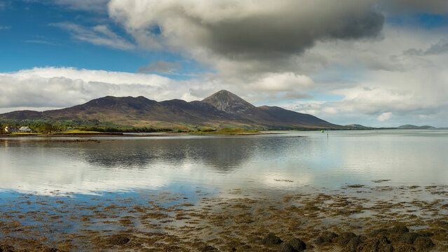 Calm ocean mirrors distant mountains under cloudy sky. Water reflects hills and clouds with still surface. Shoreline shows wetland grasses and calm ripples. Croagh Patrick. Travel Ireland scene.