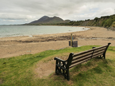 Wooden bench faces calm bay with Croagh Patrick in the background. Grassy slope meets sandy shore under cloudy sky. Old head beach in county Mayo. Travel Ireland theme. Popular landmark area.