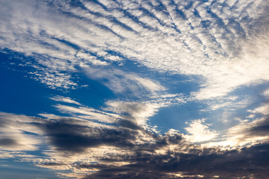 Dynamic Altocumulus clouds stretching across a deep blue sky during golden hour create a dramatic atmospheric display with soft textured formations and glowing light rays at dusk