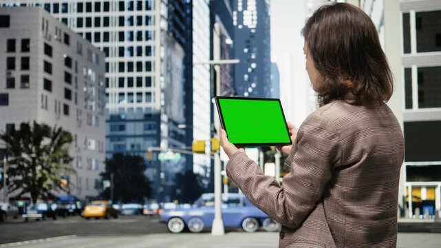 Businesswoman holds a green screen tablet in downtown and is seen from behind; the chroma key tablet mockup appears against city architecture.