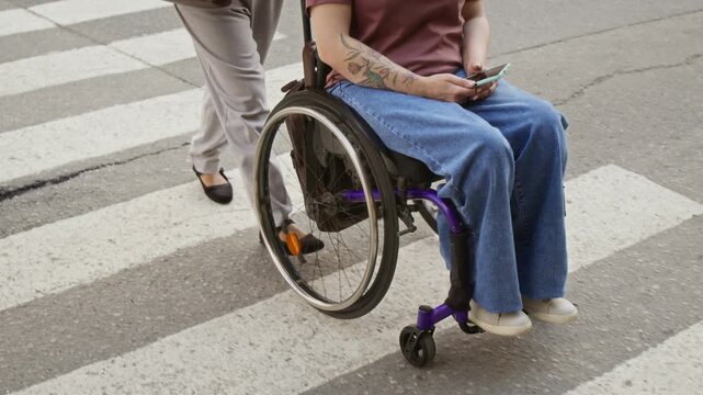 Young adult Caucasian woman with disability in wheelchair holding smartphone being pushed across crosswalk by assistant. Footage ideal for urban accessibility campaigns