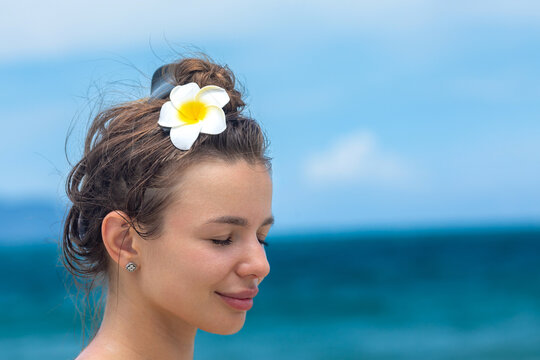 Close up head of young woman with brown hair and a white flower in her bun smiles against a tropical beach backdrop with blue ocean and sky. Side view. Copy space.