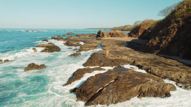 Couple standing on rocky shoreline San Juanillo beach with ocean waves splashing