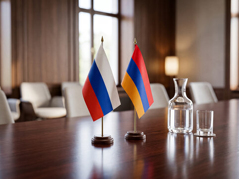 table flags of russia and armenia stand together on a polished wooden table in a meeting room near a decanter and a glass of water denoting official diplomatic talks.