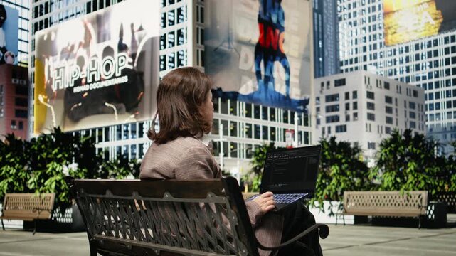 Female programmer seen from behind writes code on a laptop on a park bench; the developer uses a programming interface while working remotely outdoors.