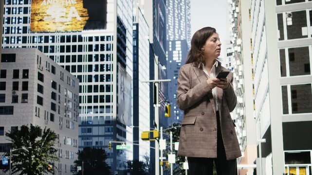 Businesswoman checks messages on smartphone while standing on sunlit downtown street; Female professional reads digital notifications on mobile phone against urban office backdrop.