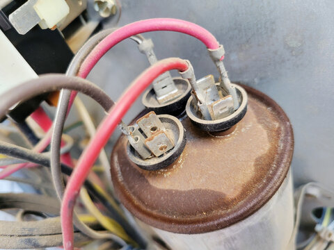 Close up view of three terminals of rusty and leaking dual run capacitor terminals connected to red and brown wires in a dusty HVAC unit