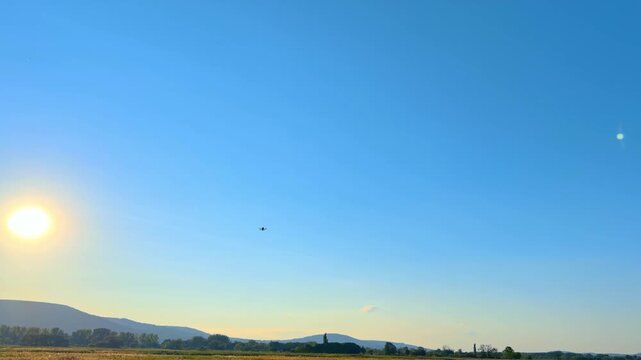 Sun shining over rural field and distant hills with flying drone. A tiny quadcopter silhouette hovers in a clear blue sky above green vegetation at golden hour.