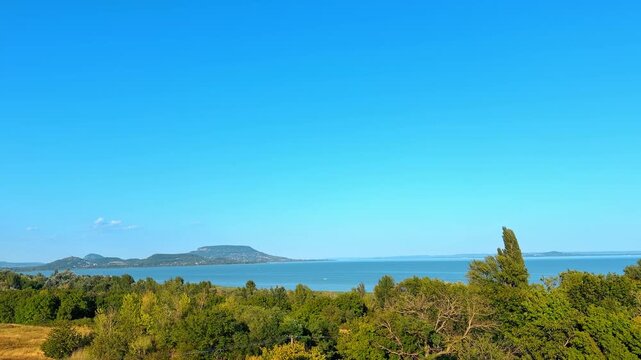 Elevated view of large blue lake surrounded by green forest. Dense green trees fill the foreground above broad calm water stretching to distant hills.