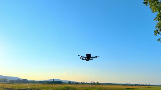 Drone flies over open field at sunset. A drone hovers above a grassy field during sunset. The sky is blue with a few clouds.