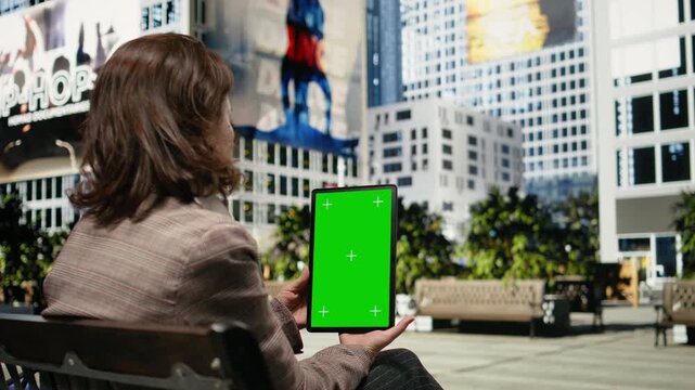 Businesswoman uses an isolated screen tablet while seated on a park bench; the mockup device is framed by urban greenery and office buildings.