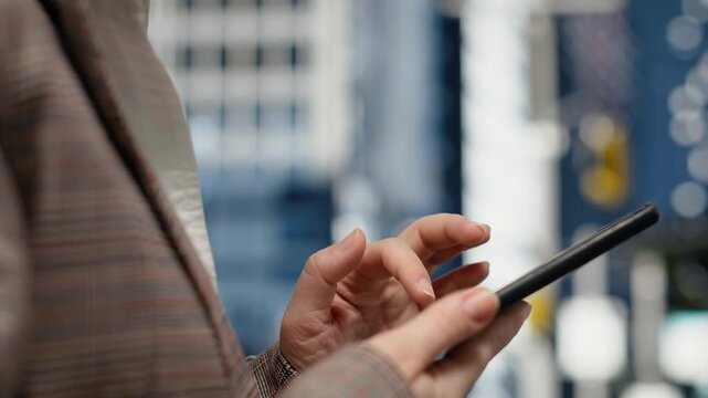 Extreme close-up view shows a smartphone in a businessperson's hands while the touchscreen is used on an urban street against a metropolitan city background.