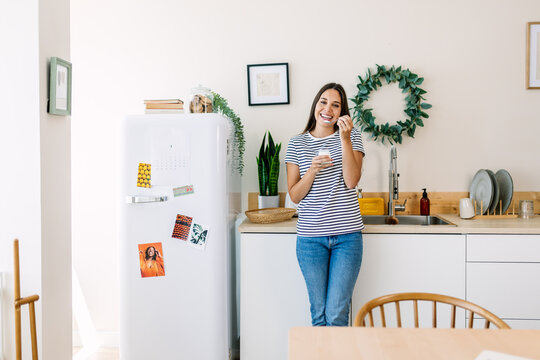 Smiling young woman enjoying yogurt at home kitchen. Happy female is eating a nutritious snack during a relaxed moment. Healthy food and lifestyle concept.
