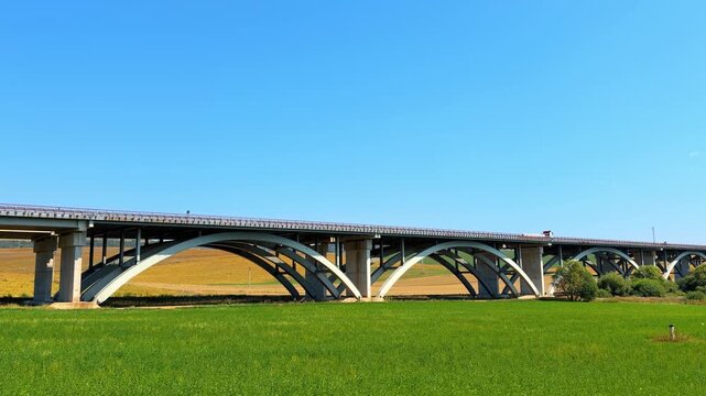 Concrete arch bridge spanning across green field. The large viaduct carries road traffic above the agricultural landscape under a clear sky.