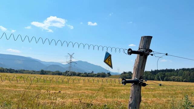 Electric fence post with warning sign in field. Transmission towers and distant mountains stand beyond the yellow high voltage symbol.