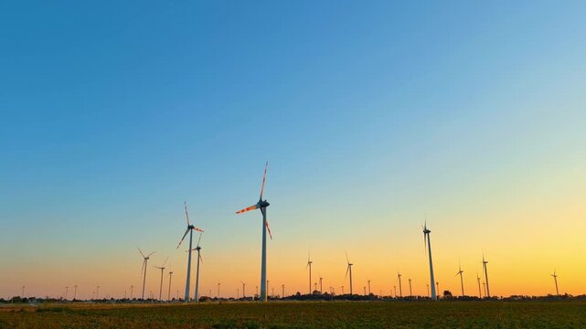 Wind turbines at sunset in an open field. Wind turbines stand tall in an open field as the sun sets in the sky. The scene shows clear blue skies with warm colors.