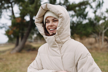 Woman outdoors wearing a beige puffer coat with hood up, smiling and enjoying crisp weather in a park setting, her joyful expression captures warmth against a chilly day with soft natural light, © SHOTPRIME STUDIO