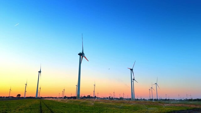 Wind turbines and field irrigation at sunset. Sprinklers water green field near tall wind turbines against colorful sunset sky.