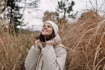 Fototapeta premium Woman in a warm coat and beanie gazes upward among tall dried grass in a winter meadow, outdoor portrait with soft natural light and a peaceful, hopeful mood.