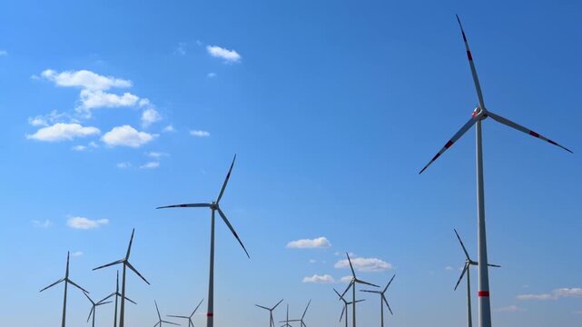 Wind turbines stand tall under blue sky. Wind turbines are arranged in rows on a clear day with blue sky and scattered clouds overhead in an open area.