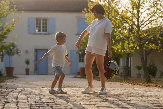 mother and child walking together on cobblestone street in residential area with blue shutters and potted plants, peaceful everyday life, eye-level perspective.