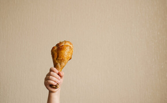 Toddler hand holding a golden roasted chicken leg drumstick against a neutral wall