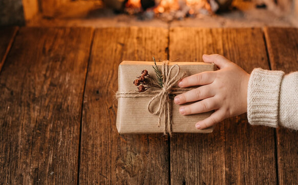 Child holding a wrapped Christmas present on a rustic wooden table by the fireplace