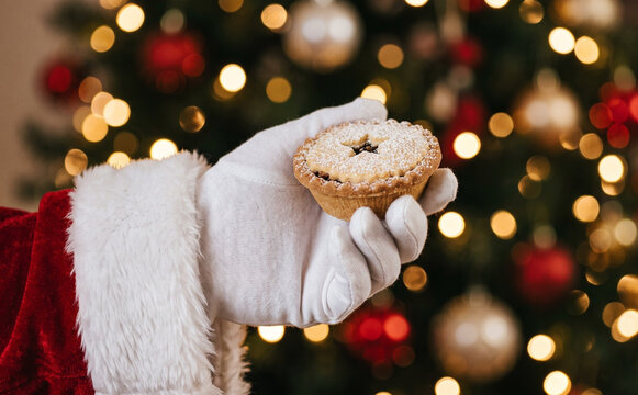 Santa Claus holding a traditional mince pie with a star cutout in front of a glowing Christmas tree