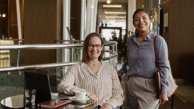 Portrait of Caucasian woman sitting with laptop and young adult Hispanic woman standing in hotel lobby, smiling. Video is ideal for corporate diversity and business travel promotions