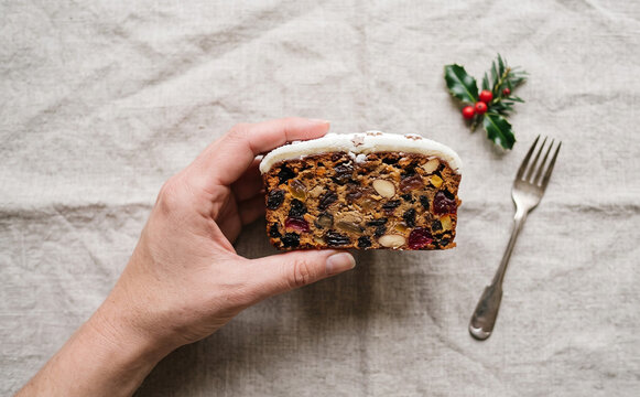 Hand holding a slice of traditional Christmas fruitcake with icing and festive holly decoration on rustic linen background