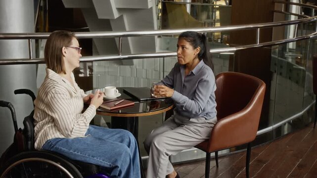 Caucasian young adult woman with disability in wheelchair and Hispanic woman discussing project and shaking hands. Perfect for corporate diversity and inclusive hiring videos