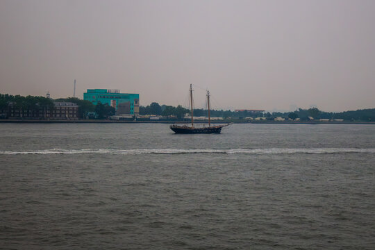 A two-masted schooner boat sailing across New York Harbor with the shoreline of Governors Island in the background.
