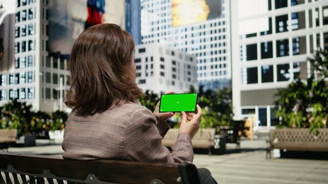 Businesswoman uses a green screen smartphone while sitting on a park bench; she checks mockup phone content during a quiet break between daily activities.