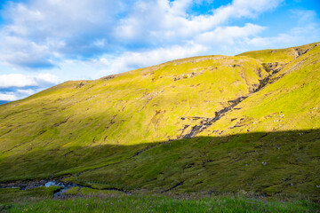 Fototapeta premium Grassy mountain slope with small streams flowing down, valley in shadow, blue sky with white clouds, Vagar, Faroe Islands, Denmark